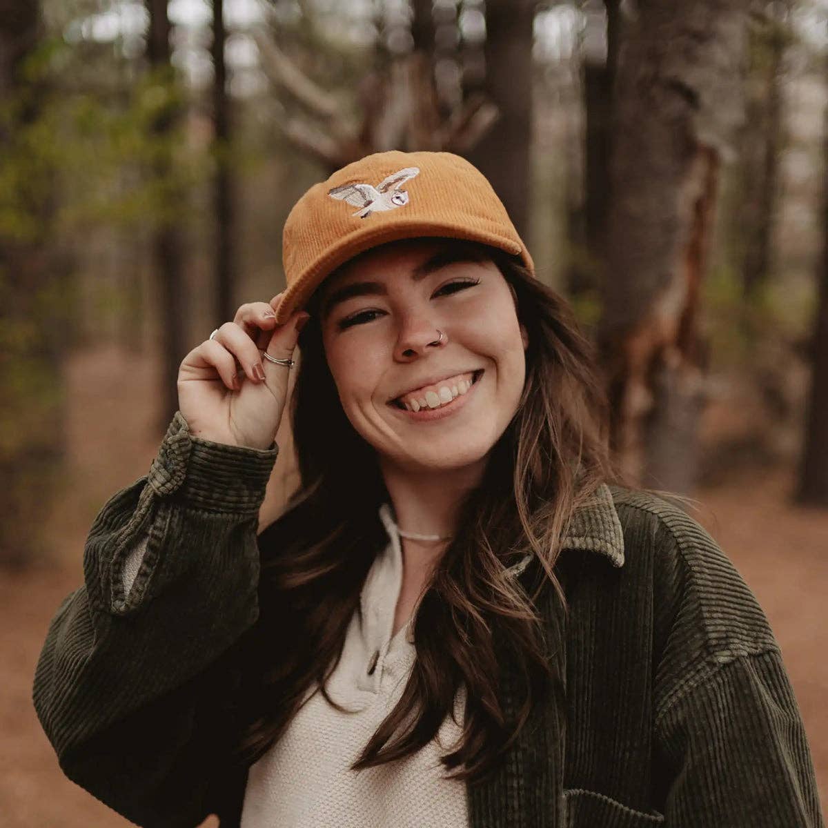 Smiling woman wears orange corduroy Barn Owl hat in a wooded outdoor setting.