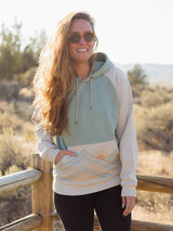 Woman wearing Rainbow Mountains Hoodie in pastel colors with a sun graphic, outdoors by a wooden railing.