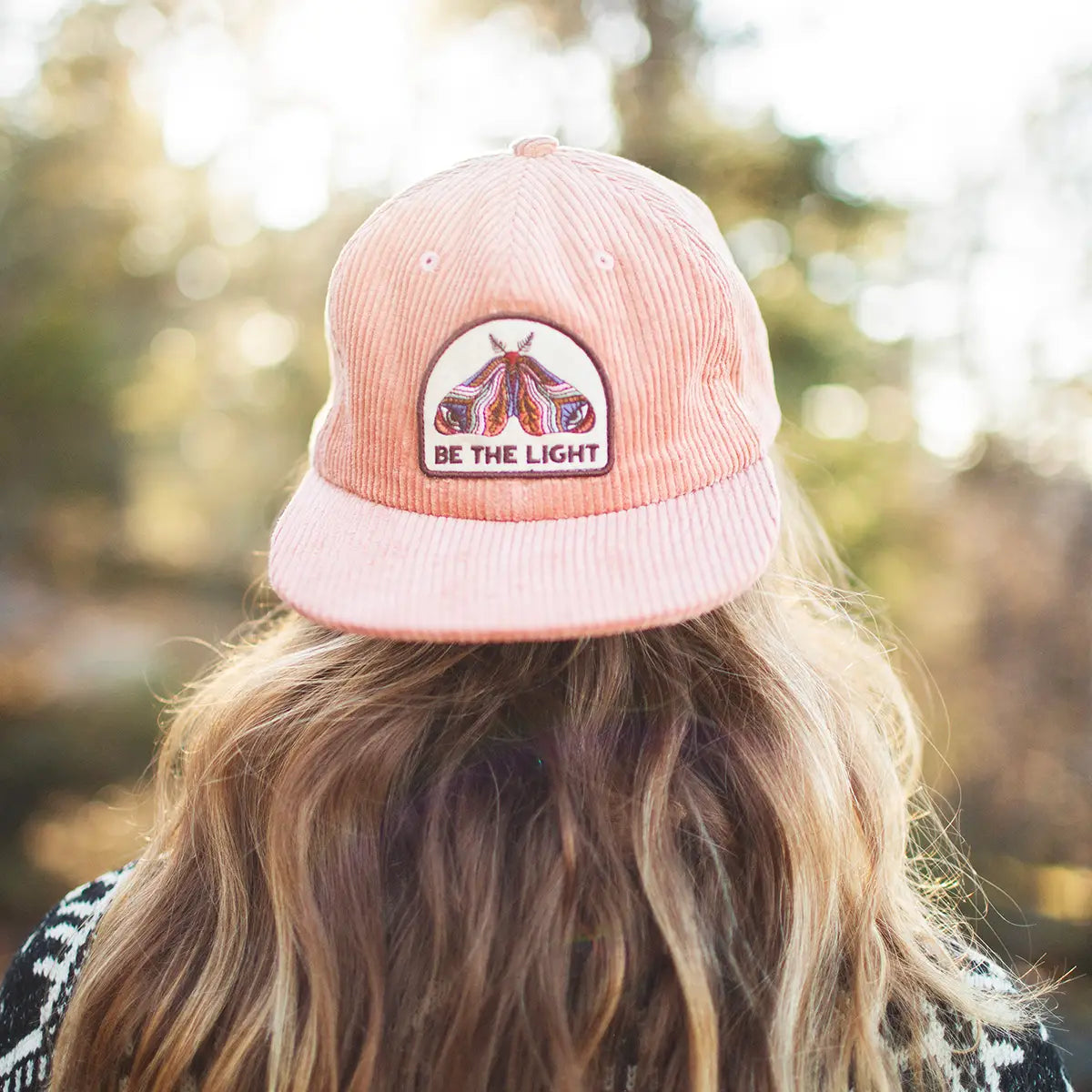 Be The Light pink corduroy flatbrim hat worn outdoors with a colorful moth patch on the front.