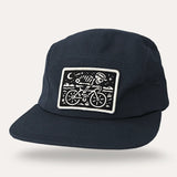 Midnight blue flat brim cap with a white bordered patch showing a skeleton riding a bicycle