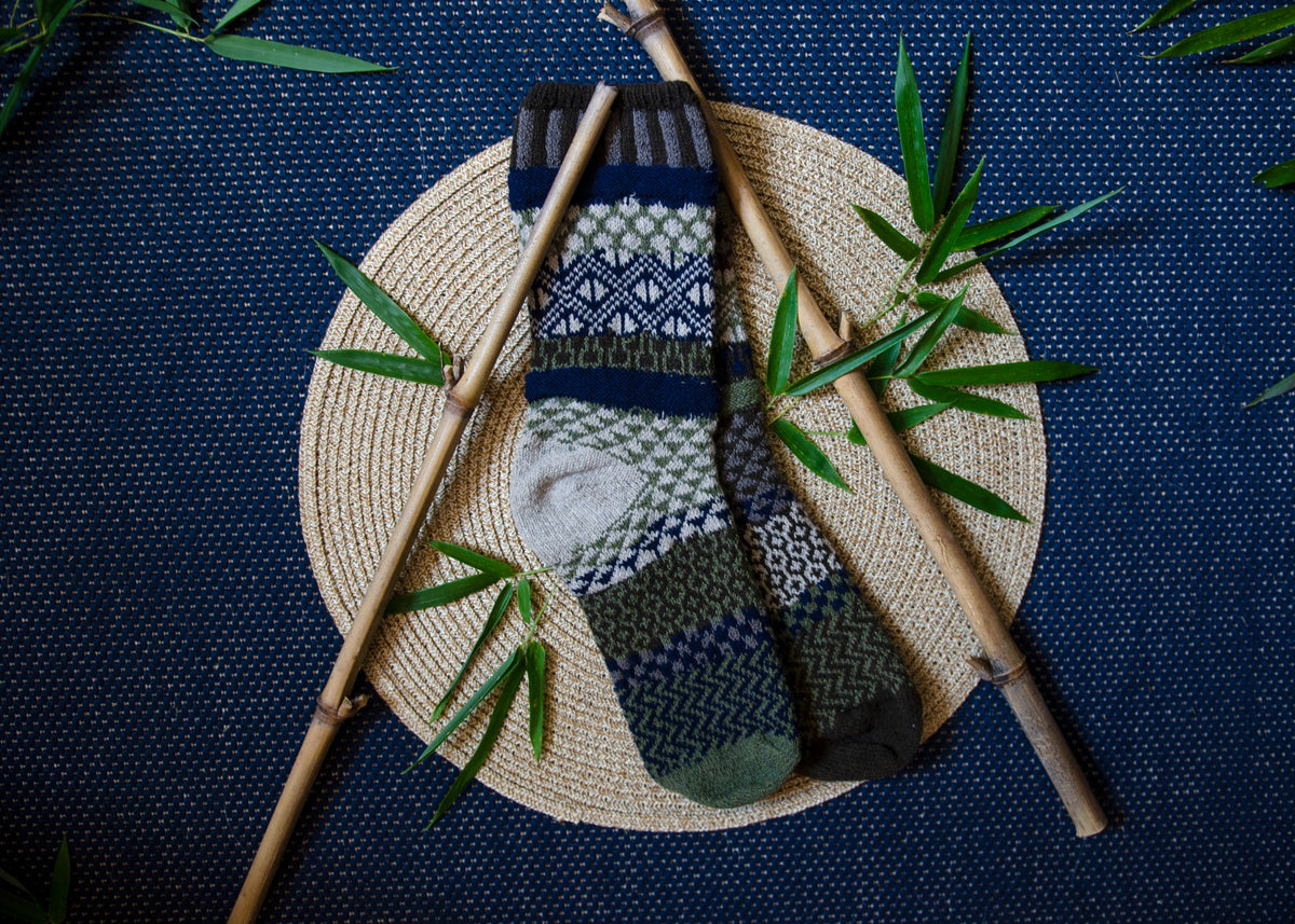 Close up of a bamboo wool crew sock featuring navy green and gray geometric patterns resting on a straw mat