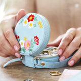 Blue round velvet jewelry case with colorful floral embroidery opened on a wooden surface with a gold ring nearby.