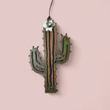 Desert cactus flower ornament in raw metal with a white bloom on top hanging from a cord against a pink backdrop