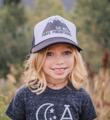 Young girl wearing a white and black trucker hat with a mountain graphic and She Will Move Mountains text