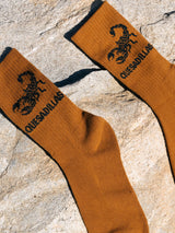 Burnt orange Quesadillas Scorpion Crew Socks with a black scorpion print on a rocky surface