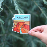 Arizona state shaped sticker showing Grand Canyon cliffs and a blue river held by a hand