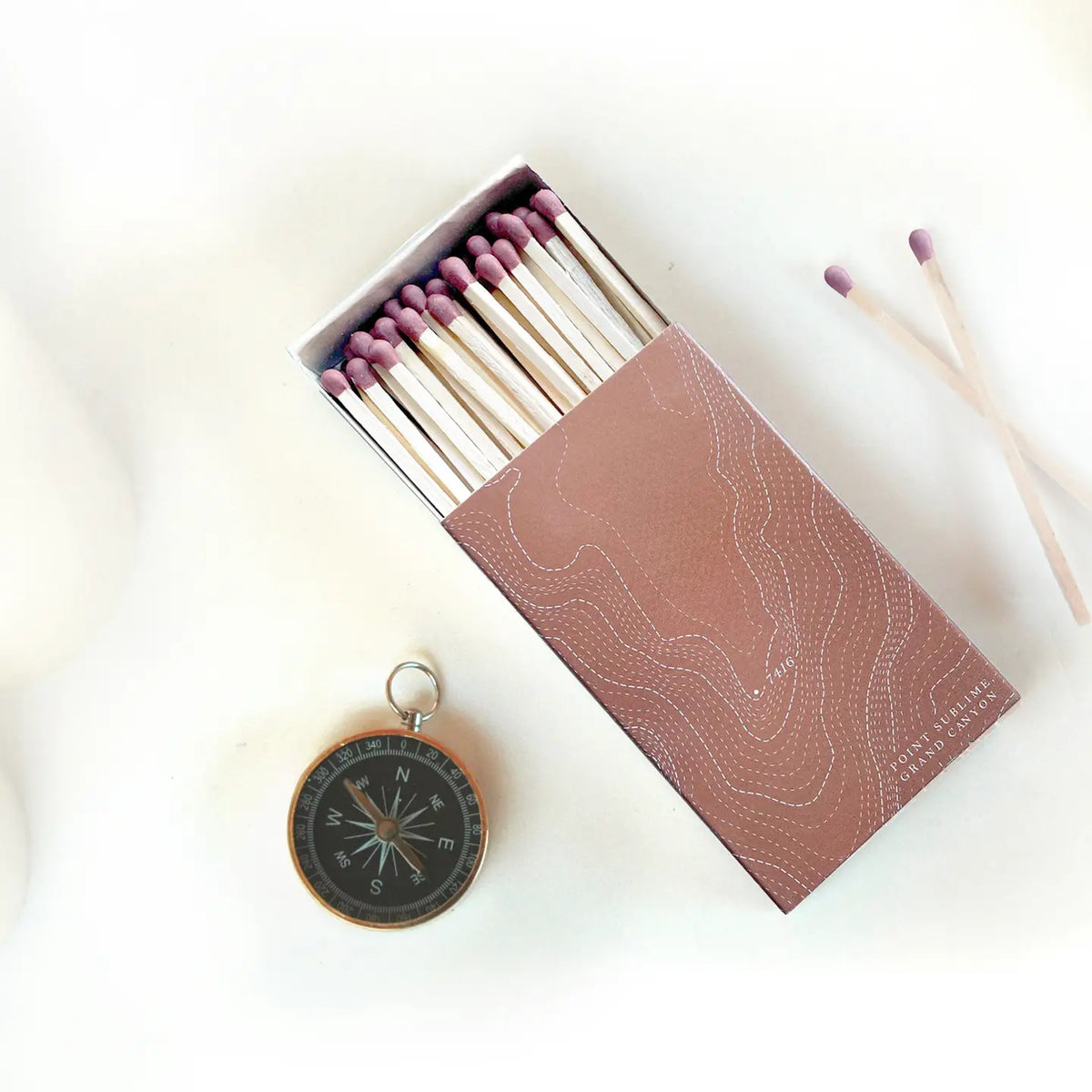 Open Grand Canyon Topography Large Matches box showing exposed matches with pink-tipped heads and a nearby compass.