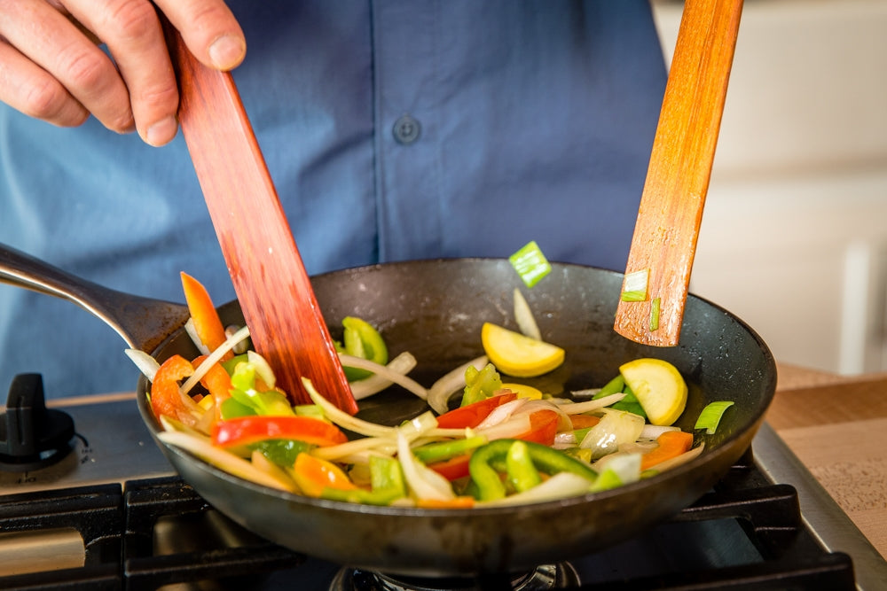 Sautéed vegetables in a black skillet being stirred with two wooden spatulas on the stove