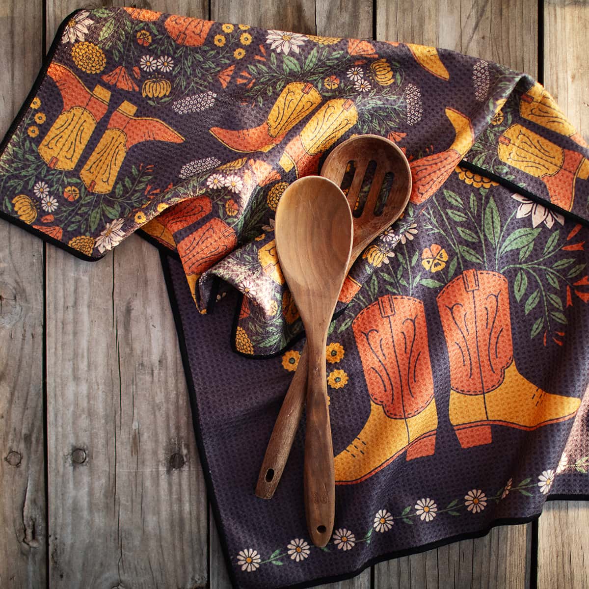 Western Bloom kitchen towel spread on rustic wood with wooden spoons featuring orange boots amid a floral print.