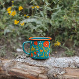 Garden Buzz enamel mug turquoise with orange rim and bold floral design resting on a log in a garden