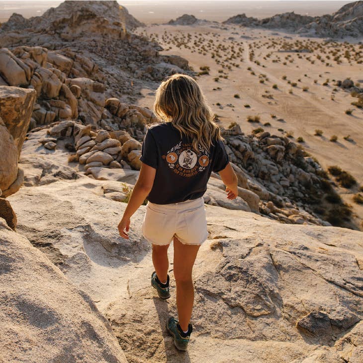 Person from behind wearing a Dead or Alive tee and light shorts on a rocky desert overlook.