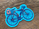 Blue bicycle sticker with brown pannier bag and gear on a wooden surface