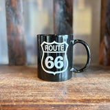 Close up of a black Route 66 mug with a white shield logo on a wooden surface.
