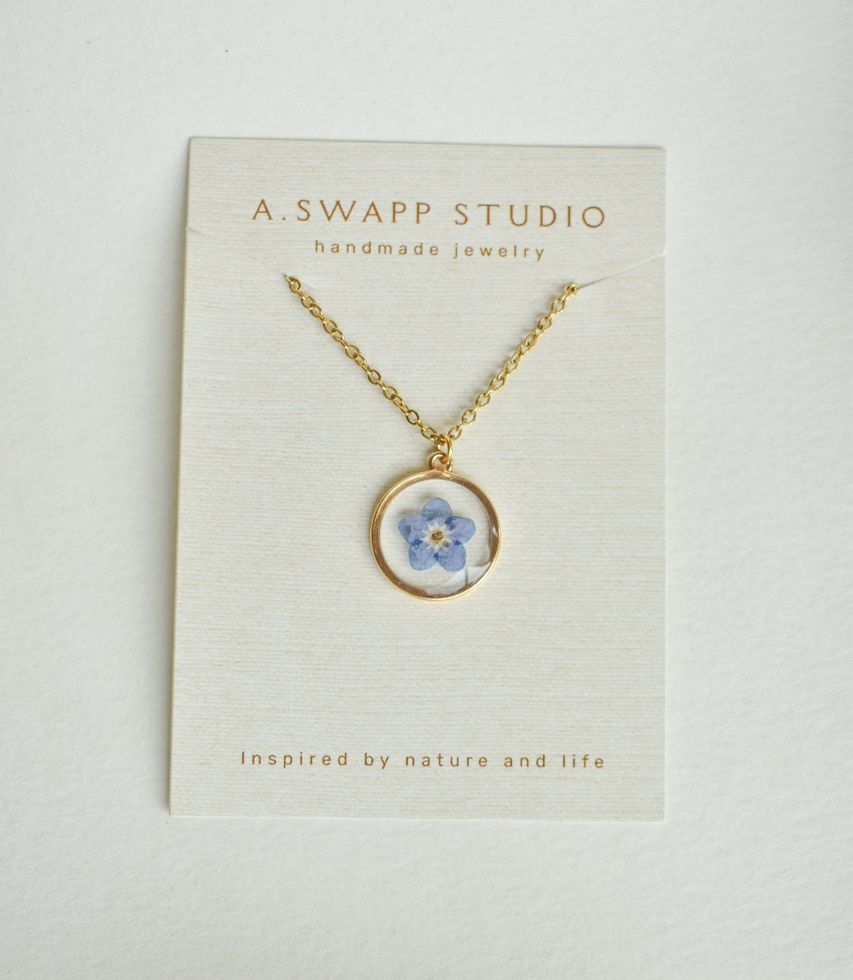 Gold circular pendant housing a blue pressed flower on a delicate gold chain against a textured jewelry card.