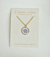Gold circular pendant housing a blue pressed flower on a delicate gold chain against a textured jewelry card.