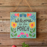 Teal wooden sign with bright floral border and the words Welcome to the Porch, hung on a rustic wood wall.