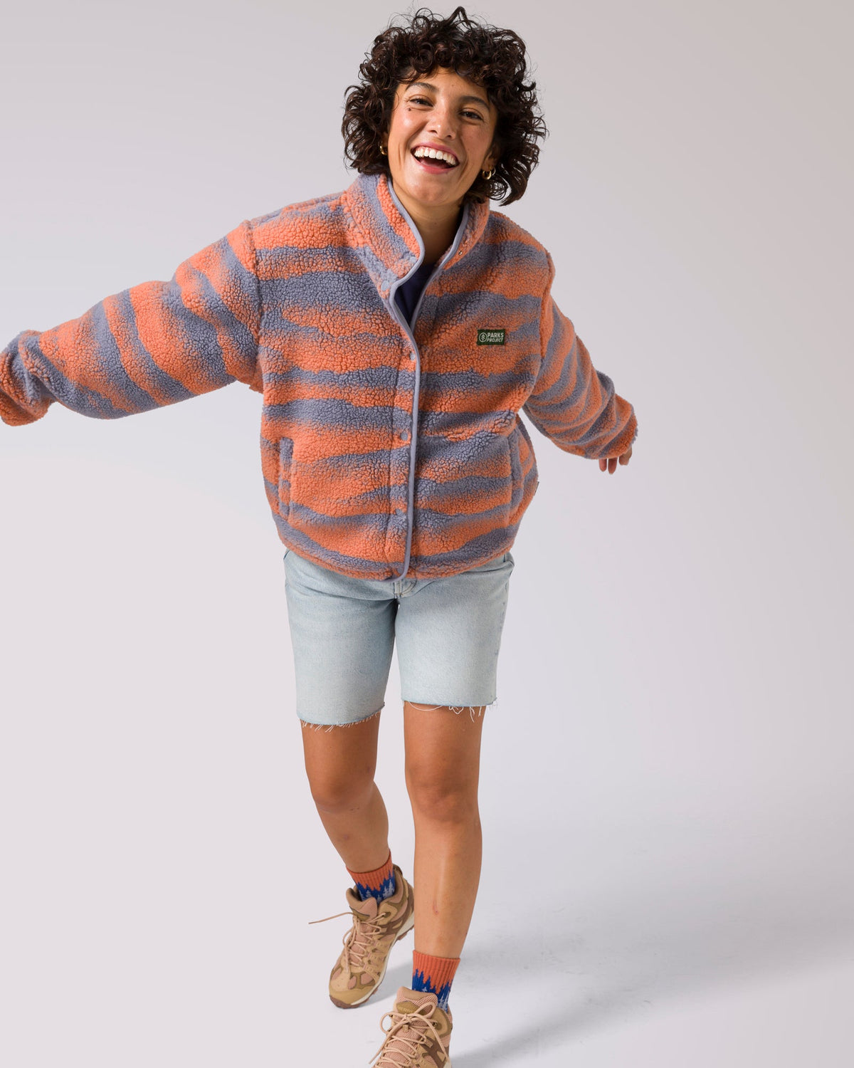 Woman wearing coral and gray striped fleece jacket with Parks Project patch, arms outstretched in a playful pose.