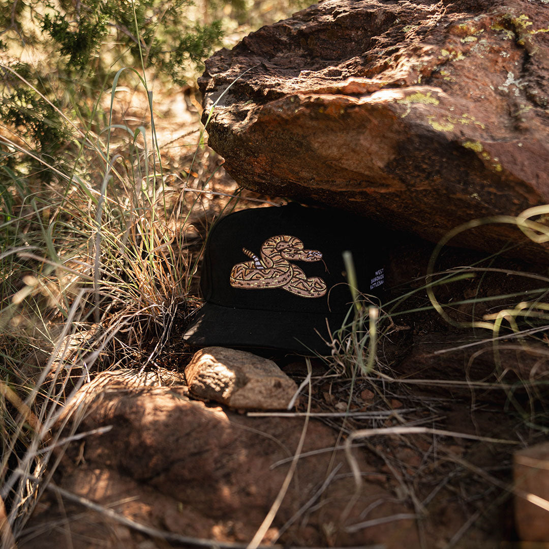 Sendero Rattlesnake Hat resting under a rock in a desert setting with the embroidered rattlesnake patch visible.
