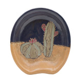 Desert motif spoon rest with pale green cacti against tan sand and a dark blue sky.