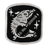 Black patch with silver embroidery of a dinosaur playing a guitar and stars.