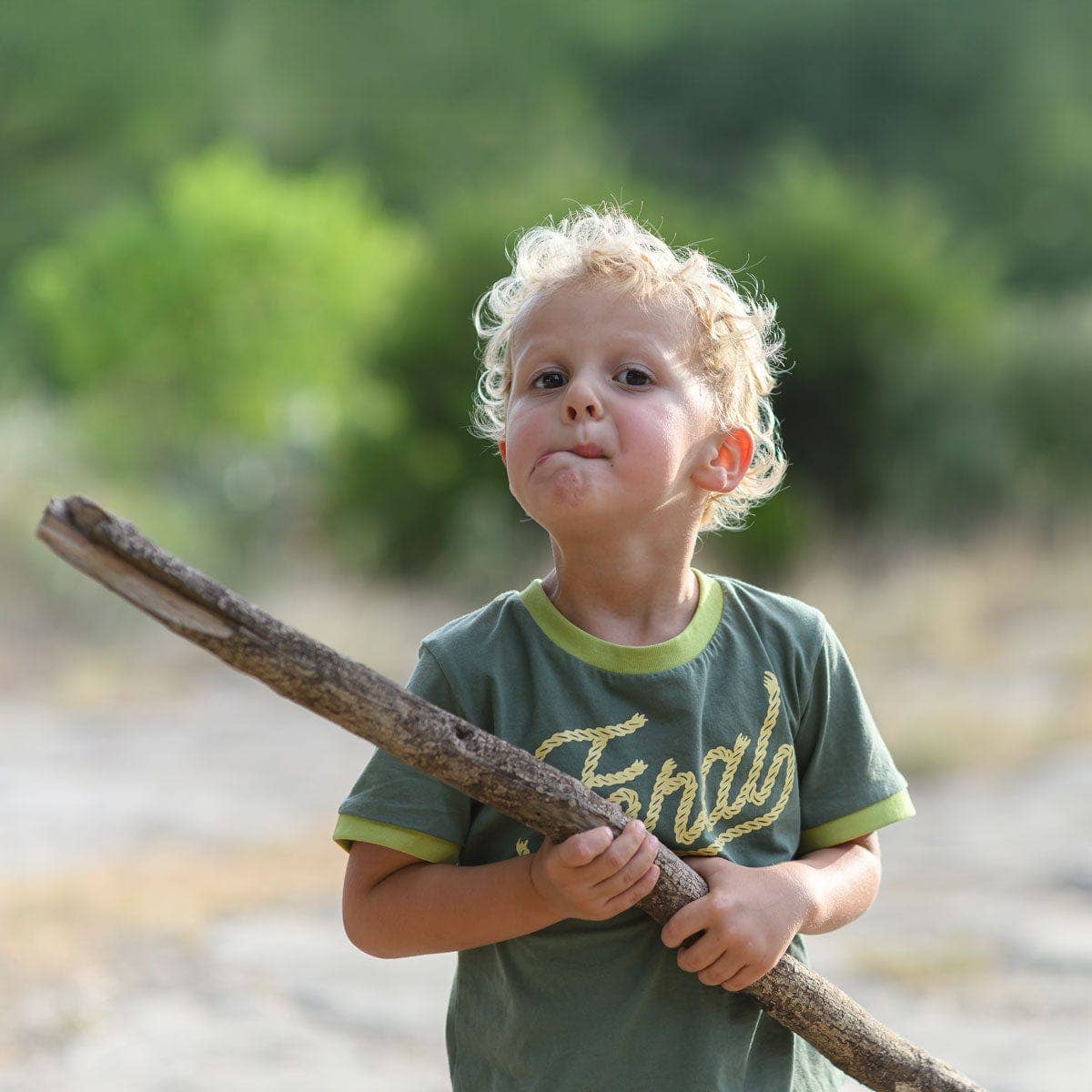 Young child wearing an olive green Feral Vintage tee outdoors, holding a long branch.