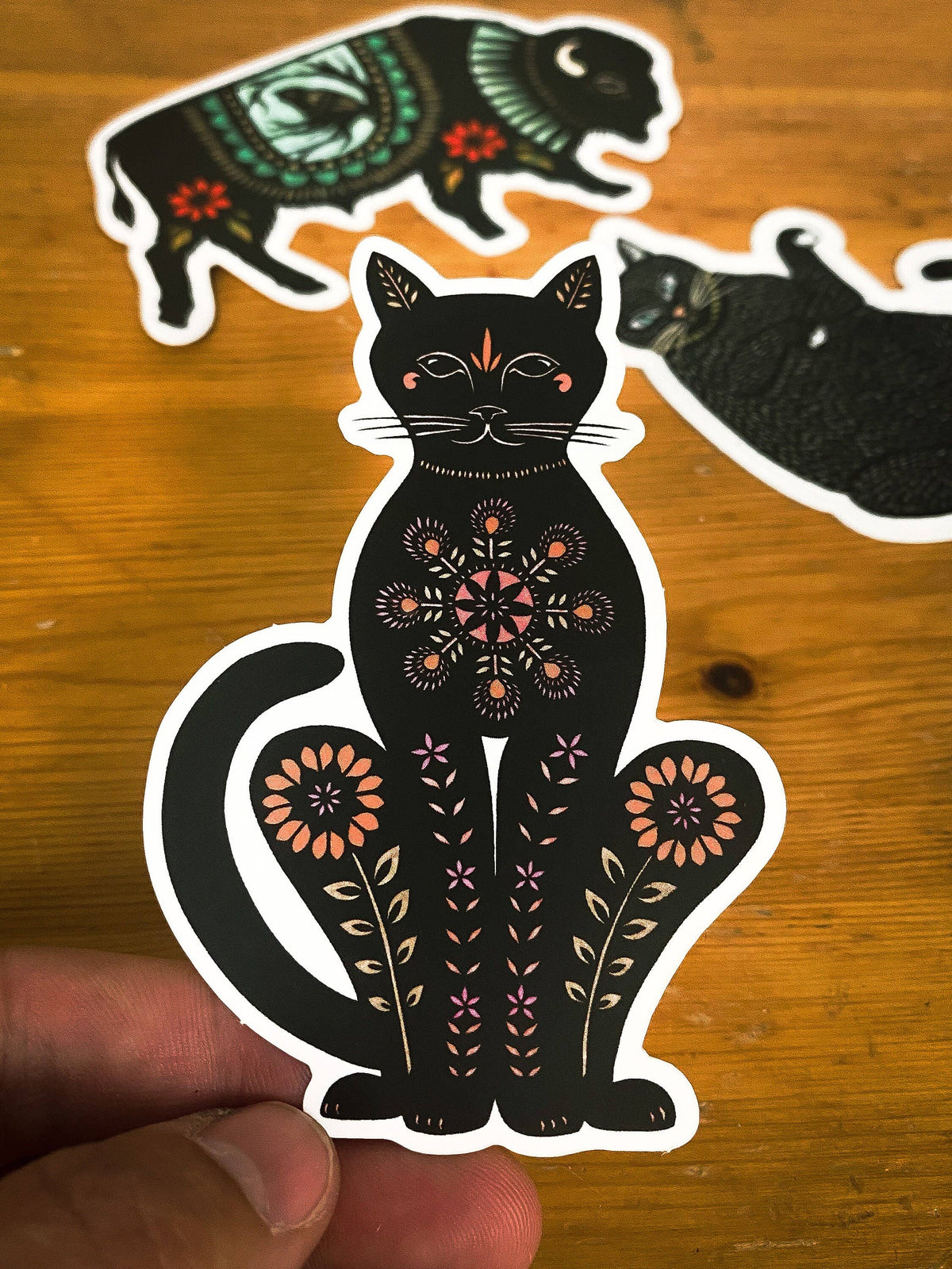 Hand holding a black cat matte vinyl sticker with intricate floral design and other cat shapes in the background
