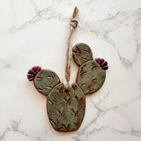 Prickly pear cactus ceramic ornament with green pads and pink flowers, suspended by twine