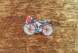 Enamel pin of a pink and blue bicycle with a red rear bag, laid on a wooden surface.