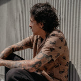 Man with tattoos wearing Brain Fried Desert button-up shirt, seated against a corrugated metal wall.