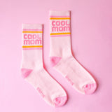 Pair of pink and white ribbed crew socks with orange and yellow stripes and Cool Mom branding on a pink background