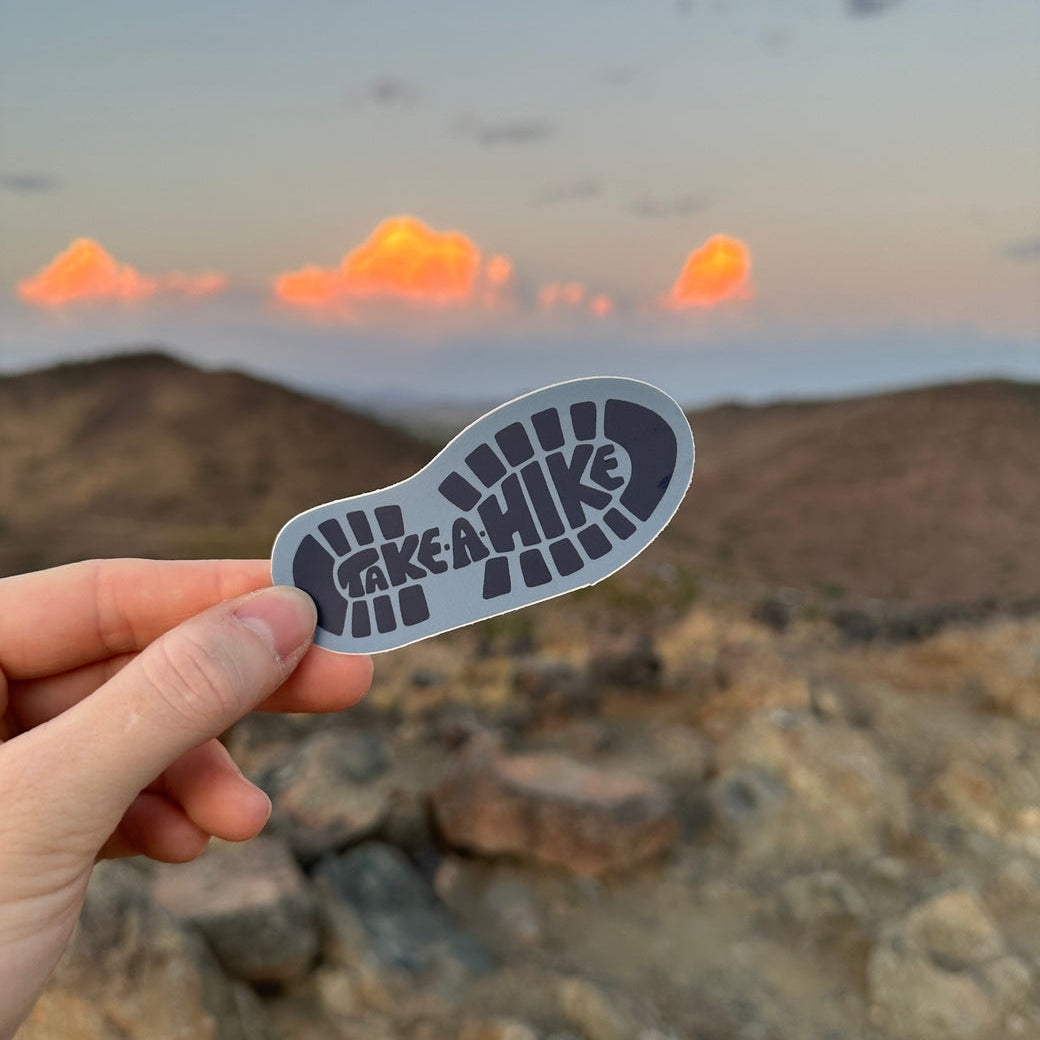 Hand holding a 'Take a Hike' sticker with a desert landscape and sunset in the background