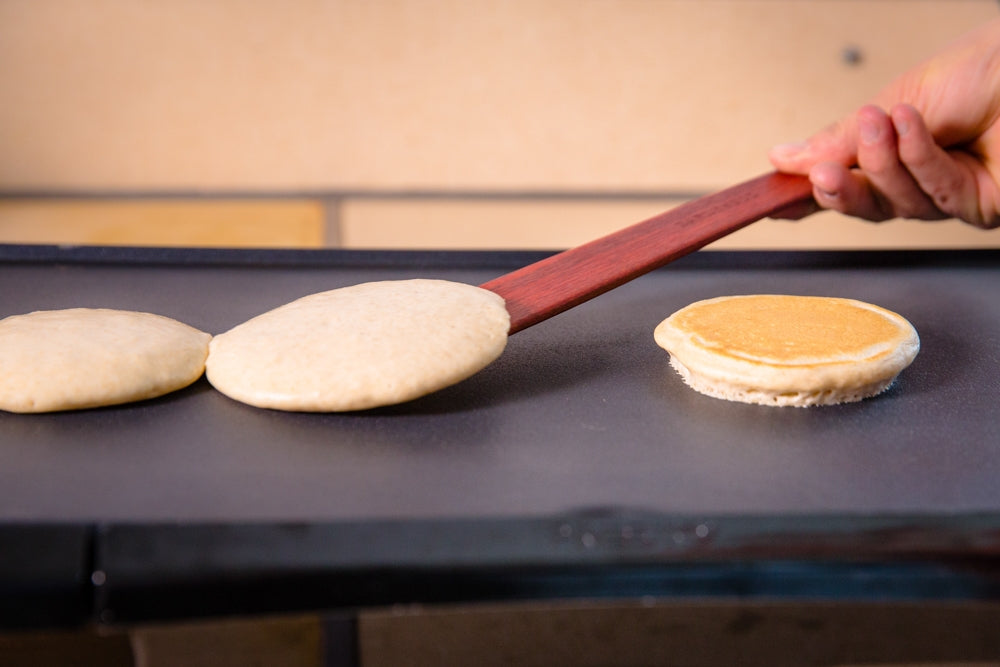 Hand using a large flat wooden saute spatula to flip pancakes on a griddle