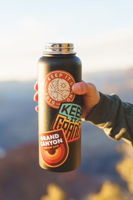Hand holds a matte black water bottle covered with Grand 1919 sticker and other outdoor decals.