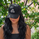 Black Daisy Dad Hat with white and yellow embroidered daisies, worn by a woman outdoors.