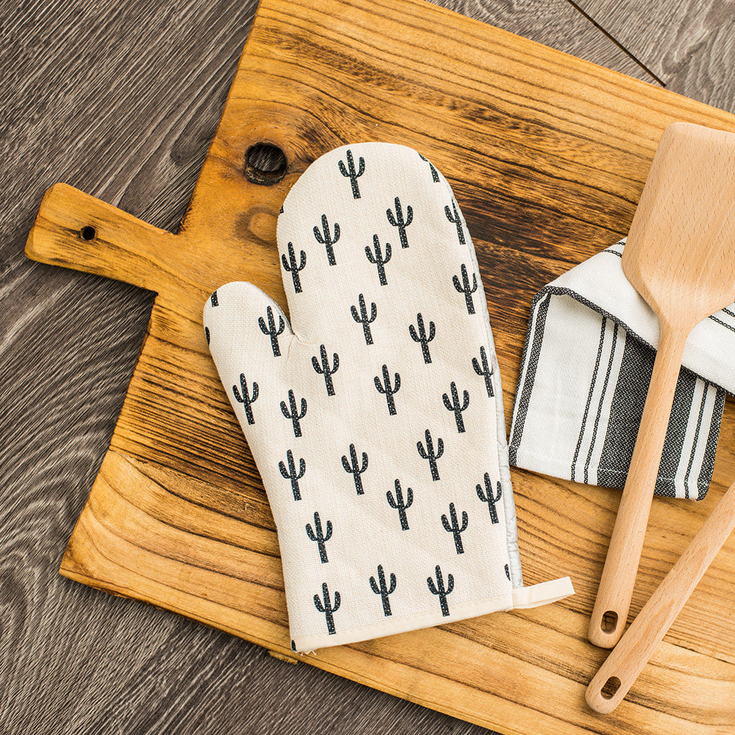 White oven mitt with a black cactus pattern resting on a wooden cutting board.