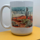White ceramic mug featuring a desert landscape with cacti and red rocks and the phrase Surrounded by Pricks