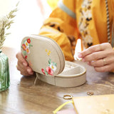 Beige velvet oval jewelry case with colorful floral embroidery opened to reveal inner compartments.