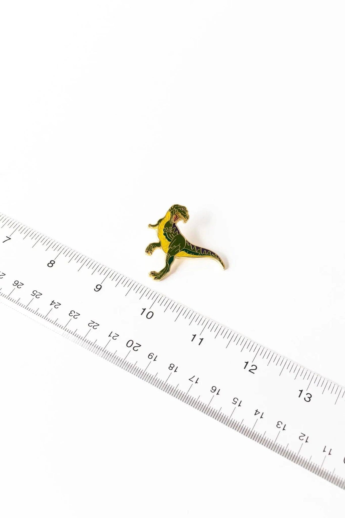 Green and yellow Tyrannosaurus Rex enamel pin next to a measuring ruler for scale