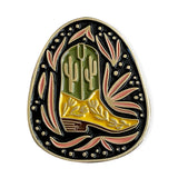 Baja Boot enamel pin close up showing a green cactus on yellow desert ground inside an oval black background with pink leaf accents and a gold edge.