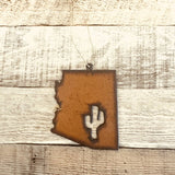Rusty Arizona state ornament with a cutout saguaro cactus hanging on twine against weathered wood