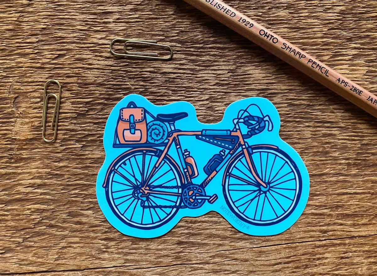 Blue bicycle sticker on a wooden surface with metal paperclips and a pencil nearby