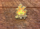 Campfire enamel pin with orange flames and logs on a wooden surface
