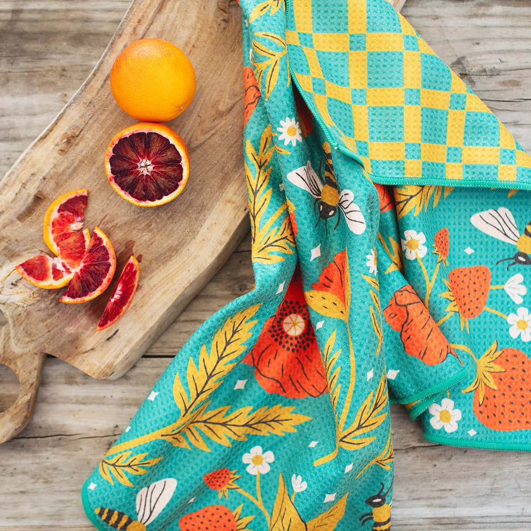 Colorful turquoise kitchen towel with fruit and bee motifs draped over a wooden cutting board beside sliced oranges.
