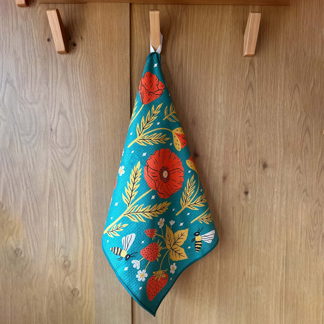 Garden Buzz kitchen towel hanging on a wooden cabinet with teal background red flowers and bees