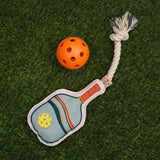 Paddle shaped rope dog toy resembling a pickleball paddle with a small orange ball nearby on grass.