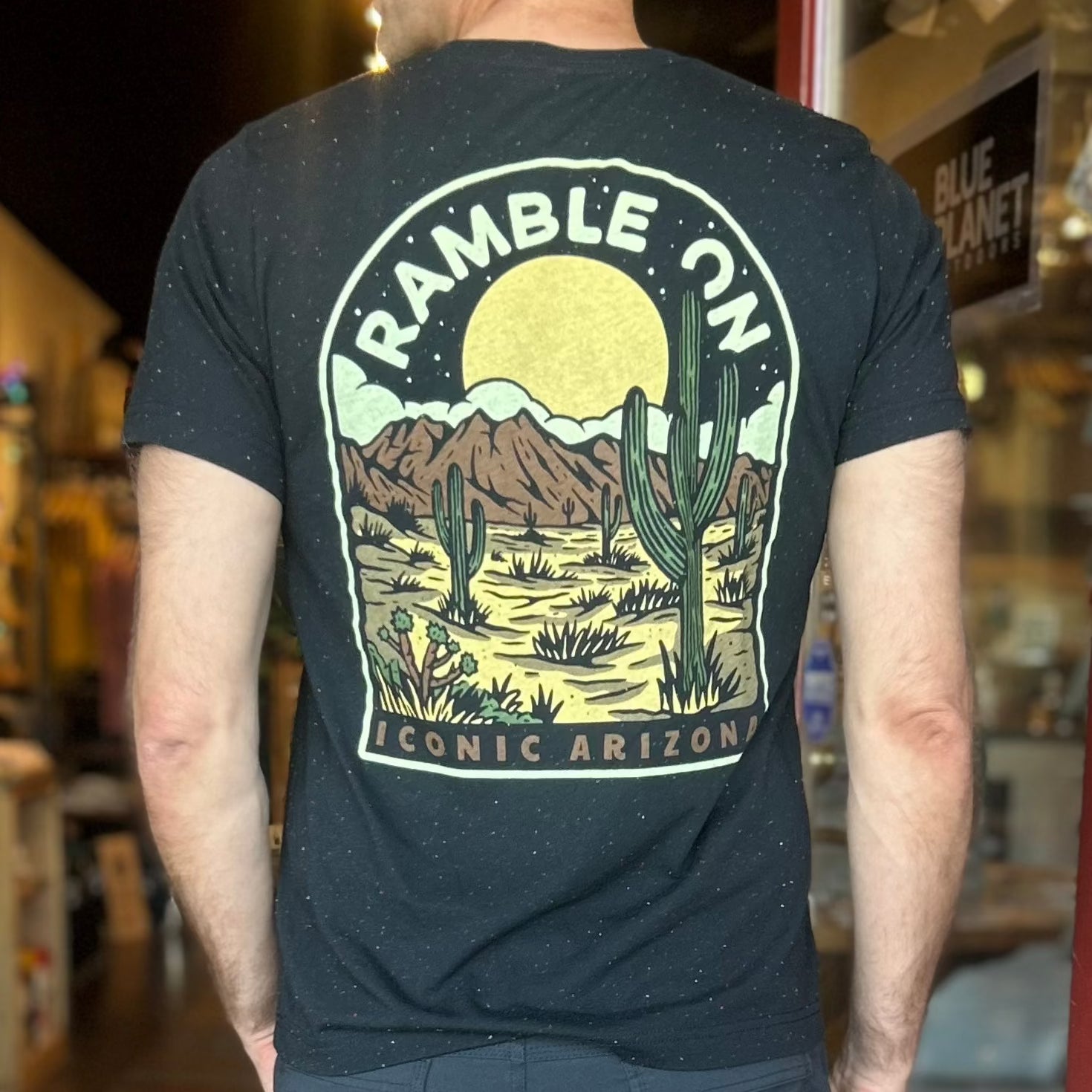 Back view of a dark speckled tee with a large circular desert scene showing sun mountains and cacti and Iconic Arizona text