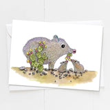 Javelina mother with two babies in a desert scene with flowering cactus plants.