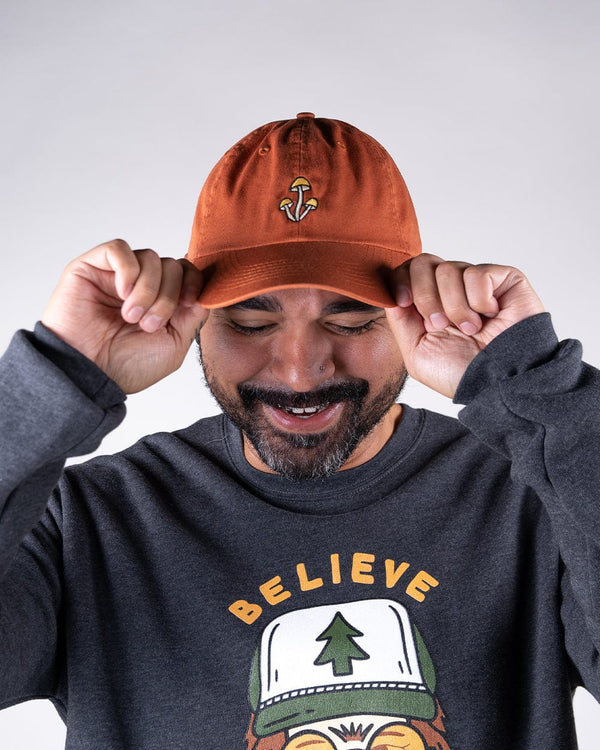 Man wearing an orange Shroom Dad Hat adjusts the brim with embroidered mushroom logo visible.