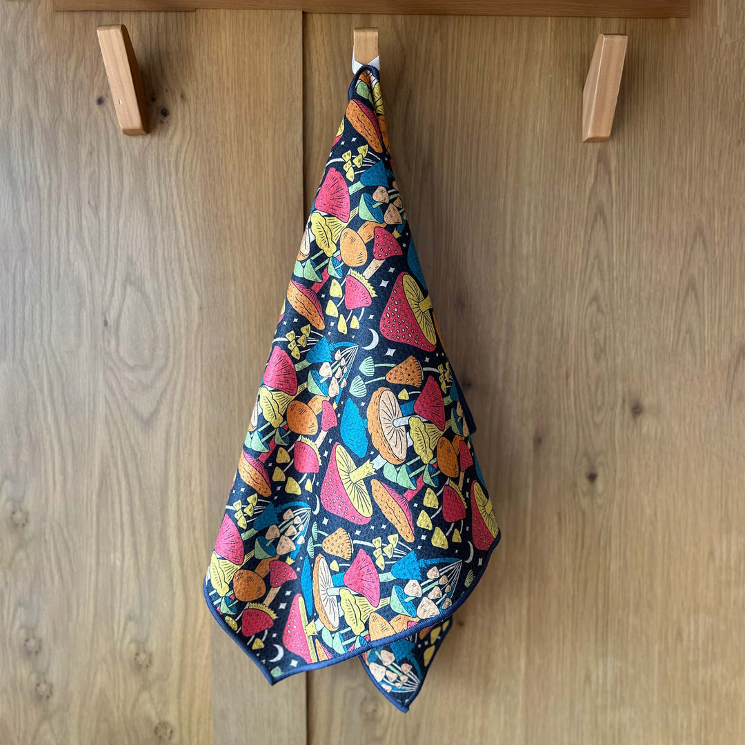 Midnight Mushroom kitchen towel hanging from a wooden hook with a vibrant mushroom print on navy fabric