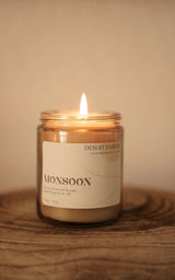 Monsoon 7.5 oz candle in a clear glass jar with a lit flame on a wooden surface.